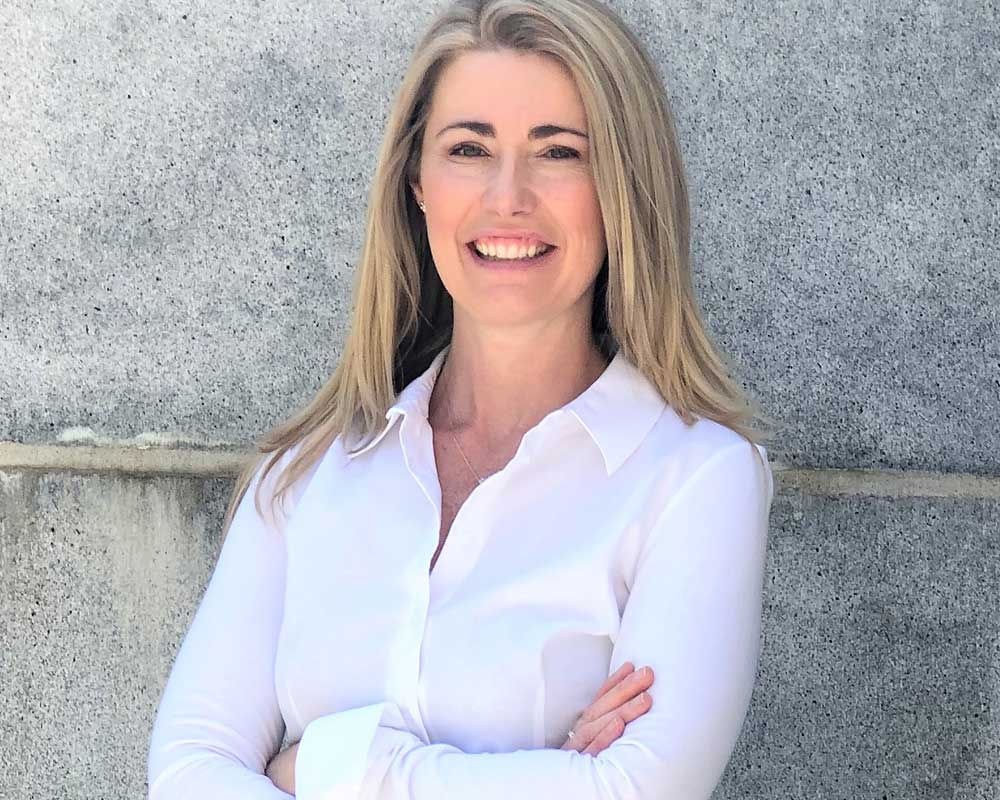 smiling woman with long blonde hair standing against concrete wall wearing white shirt with arms crossed