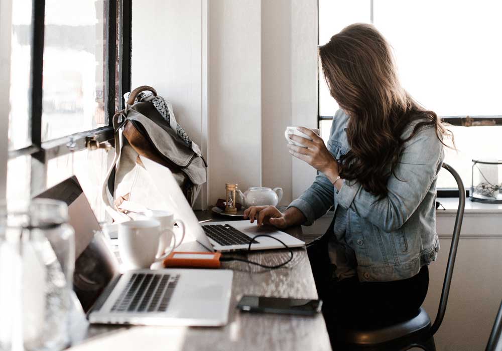 woman with long brown hair holding tea cup while sitting at desk in front of laptop